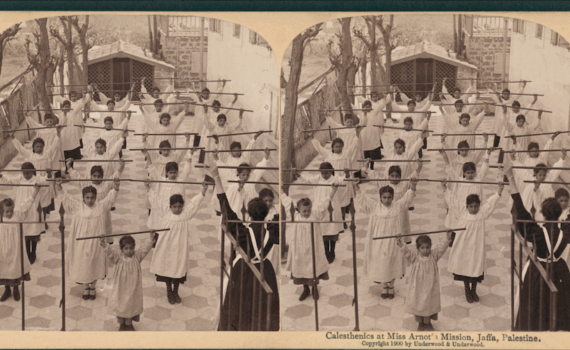 Stereoscope view of children lined up four across and six rows, with one younger children in the front, holding straight sticks over their heads. A teacher, with back turned to camera, holds a stick over their head. A caption reads "Calesthenics at Miss Arnot's Mission, Jaffa, Palestine. Copyright 1900 by Underwood & Underwood."