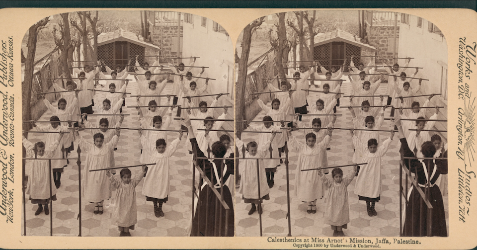 Stereoscope view of children lined up four across and six rows, with one younger children in the front, holding straight sticks over their heads. A teacher, with back turned to camera, holds a stick over their head. A caption reads "Calesthenics at Miss Arnot's Mission, Jaffa, Palestine. Copyright 1900 by Underwood & Underwood."