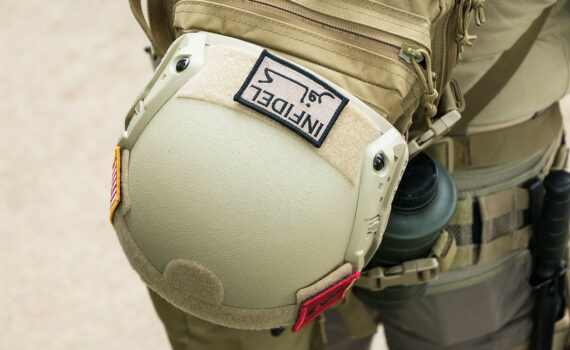 Close-up of a tan tactical helmet attached to the outside of a matching backpack. The helmet has a rectangular patch with the text “INDEL” and a symbol, plus smaller patches including a U.S. flag. The backpack features straps, compartments, and attached gear such as a water bottle. The scene is tightly framed, focusing on the gear’s texture, stitching, and muted earth-tone colors.